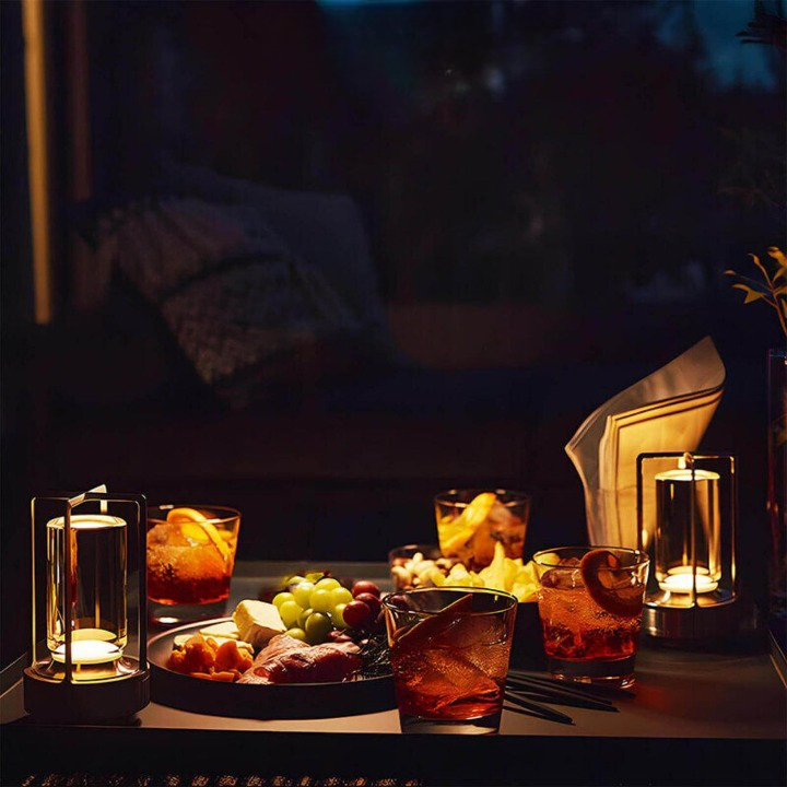 Two lamps cast warm light over an evening table of cocktails, a snack platter and a folded napkin.