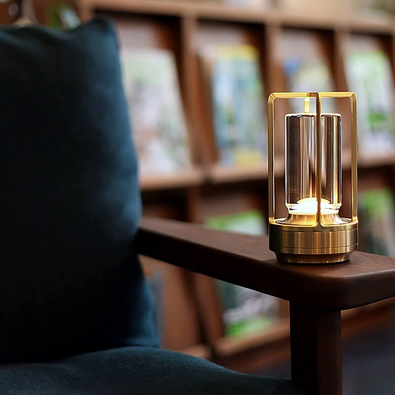 Gold-framed lamp glowing on the wooden arm of a chair, with a teal cushion and bookshelves blurred behind.