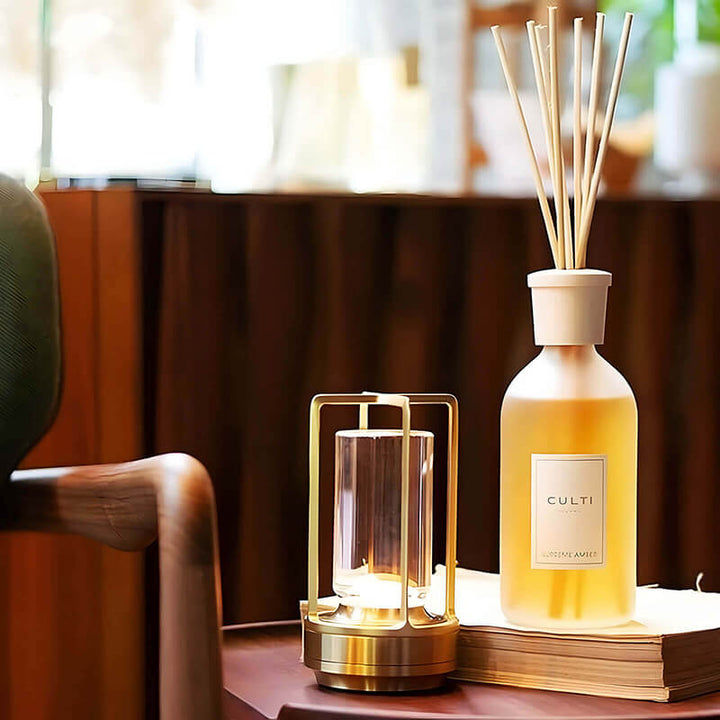 Gold-framed lamp lit on a small table next to a reed diffuser bottle and a stack of books.