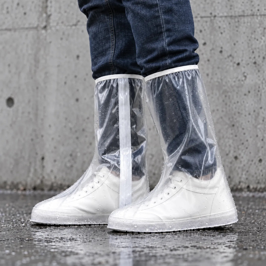 Side view of clear tall overshoes dotted with raindrops, worn over white sneakers on wet pavement in front of a concrete wall. 3