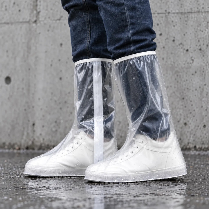 Side view of clear tall overshoes dotted with raindrops, worn over white sneakers on wet pavement in front of a concrete wall. 3