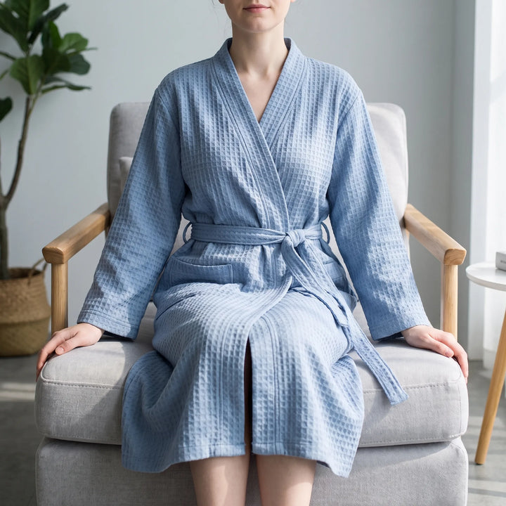 Woman seated in a light armchair wearing the blue waffle-weave robe, belt tied at the waist, in a bright living room. 1