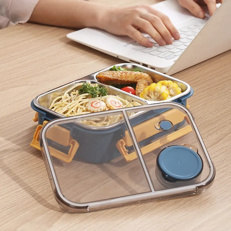 another view of navy and yellow container with noodles, chicken wing and corn on a wooden desk, clear lid in front, person typing on a laptop.