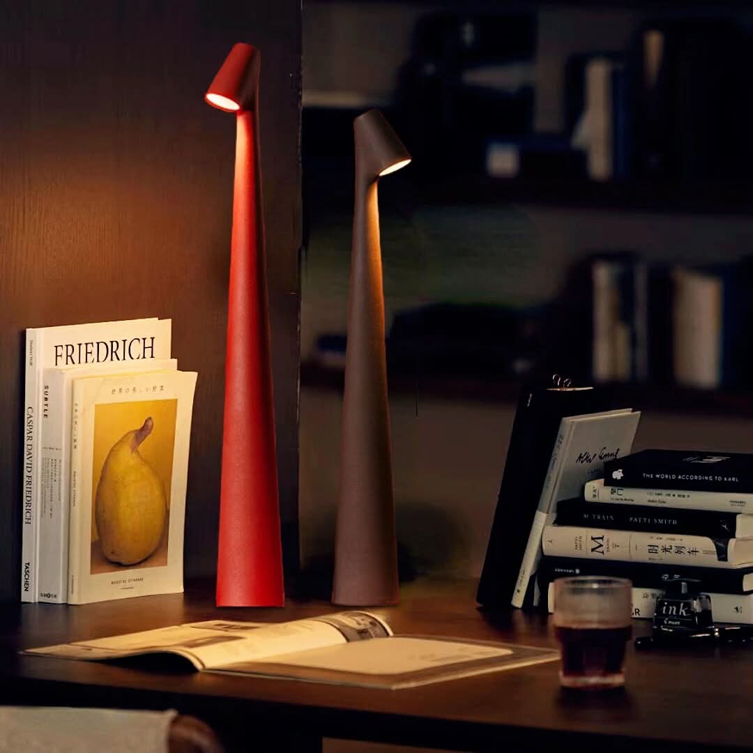 Red and dark brown lamps glowing on a wooden desk beside stacked art books, an open magazine and a glass of tea. 1