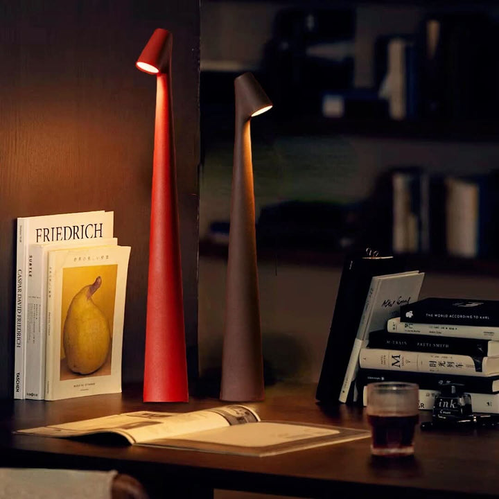 Red and dark brown lamps glowing on a wooden desk beside stacked art books, an open magazine and a glass of tea. 1