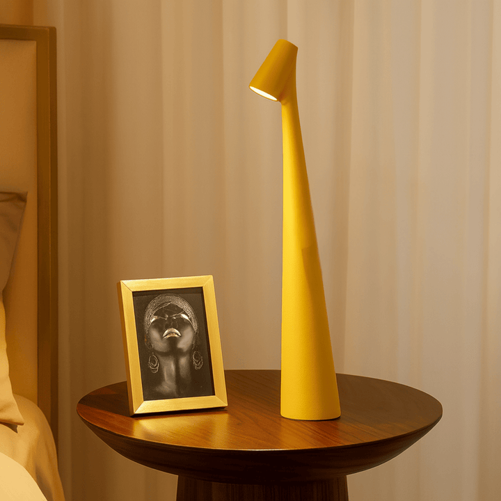 Red cordless table lamp lit on a round wooden bedside table beside a small gold-framed portrait.