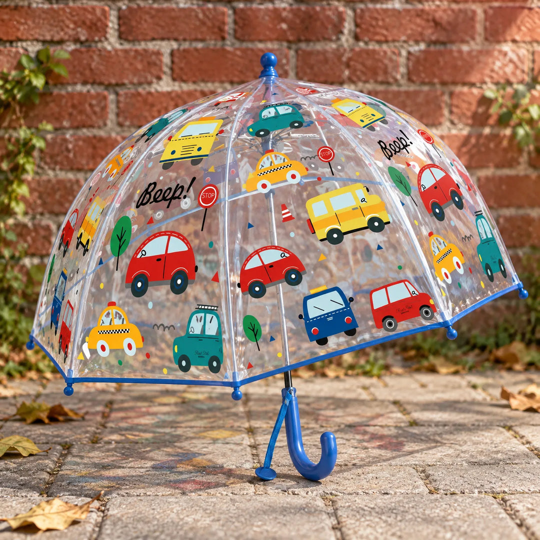 Clear dome umbrella with a blue trim and hook handle, printed with colorful cartoon cars and trees, standing outdoors by a brick wall. 3
