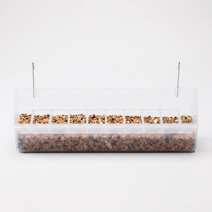 Long clear plastic feeder with two metal hooks, filled with mixed seed and a row of small divided slots along the front. 1