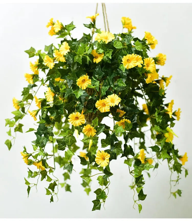 Yellow faux morning glory blooms and trailing green leaves in a hanging basket held by rope against a plain white background.