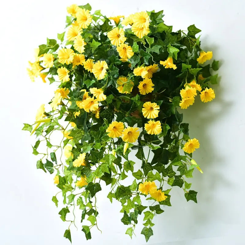 Close view of yellow faux morning glory flowers and dark green leaves with trailing vines against a white wall.