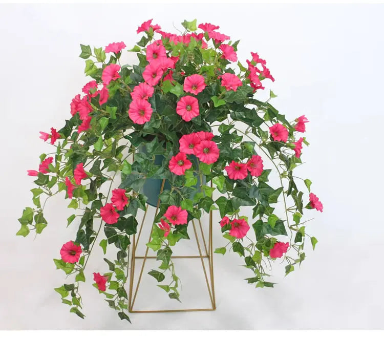 Pink faux morning glory blooms and green leaves cascading from a dark pot on a gold geometric metal stand.