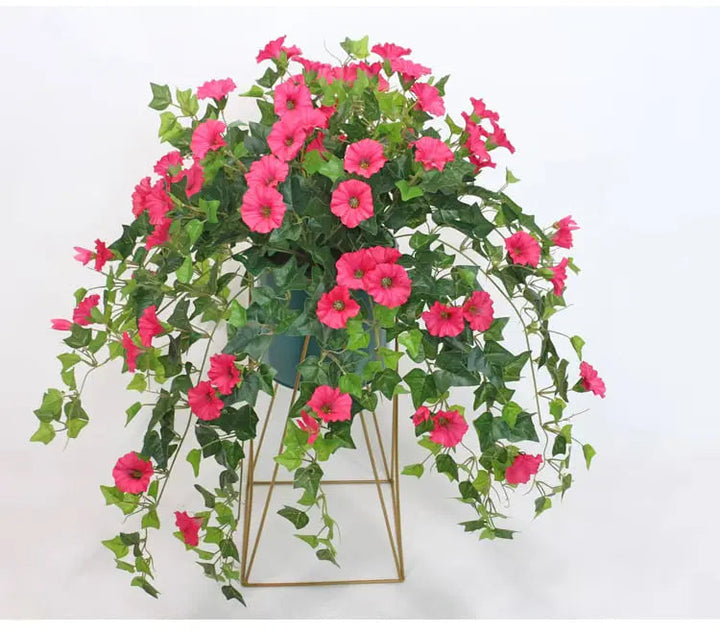 Pink faux morning glory blooms and green leaves cascading from a dark pot on a gold geometric metal stand.