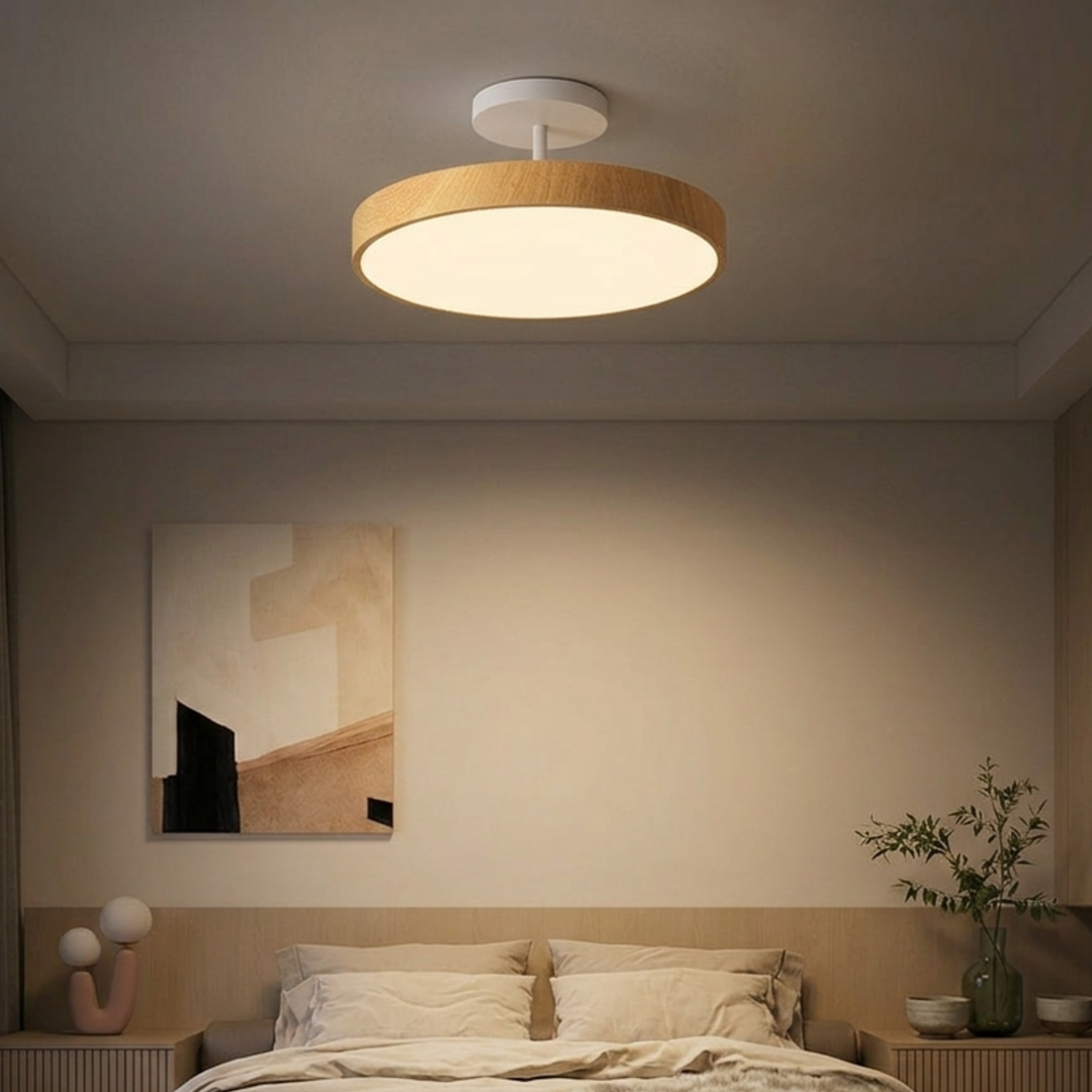 Light wood-look disc ceiling light with a white stem hangs over a bed in a calm, warmly lit bedroom.