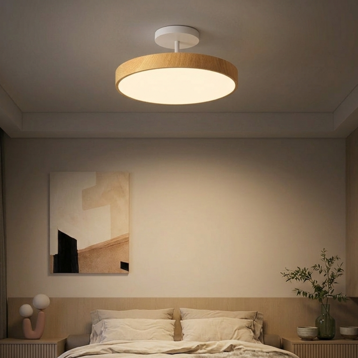 Light wood-look disc ceiling light with a white stem hangs over a bed in a calm, warmly lit bedroom.