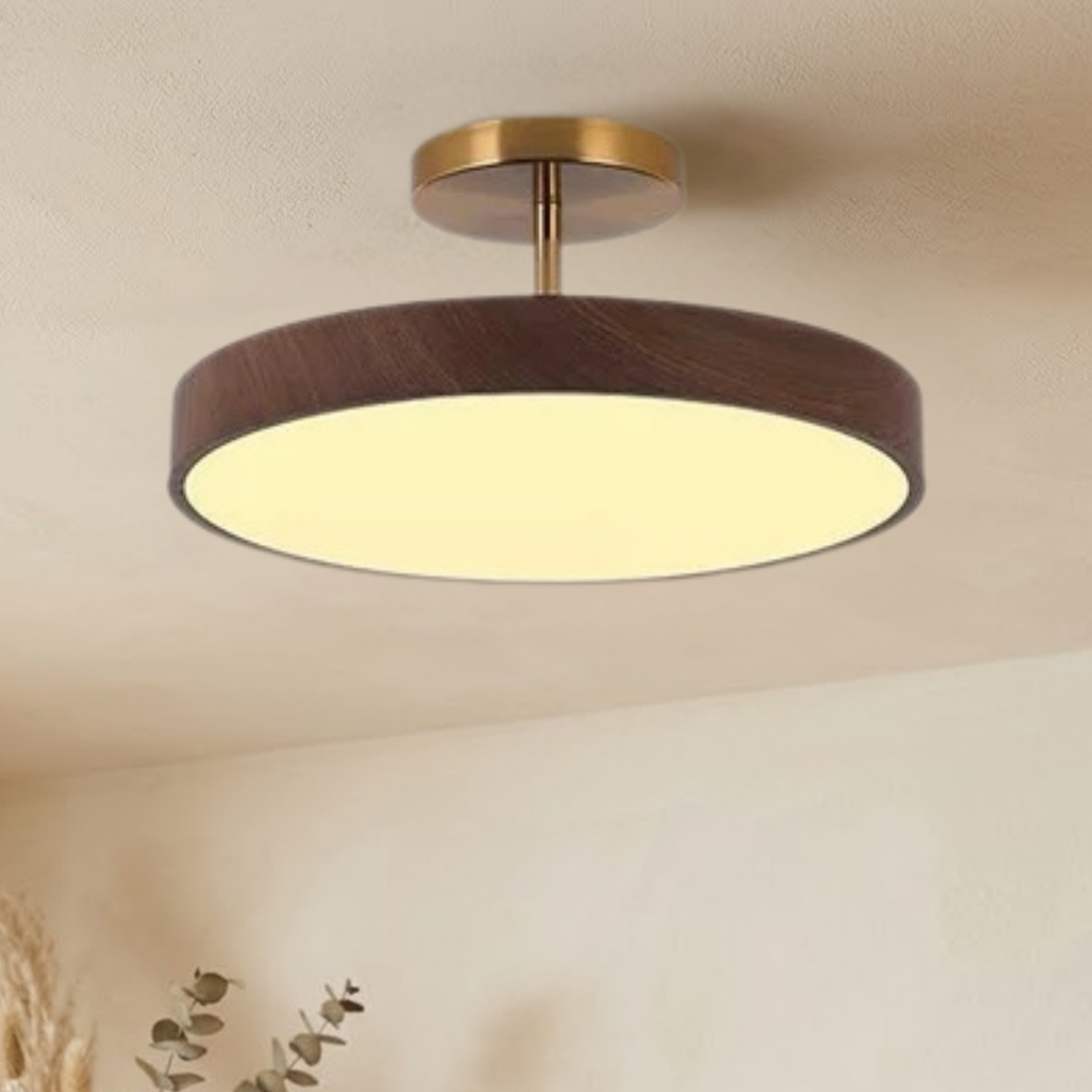 Dark wood-look disc ceiling light with brass-tone stem, seen from slightly below and glowing warm against a beige ceiling.