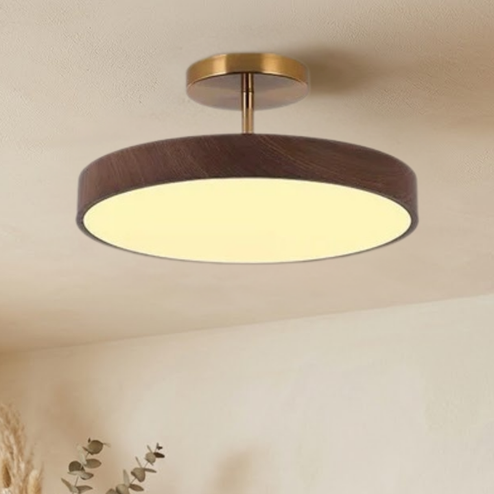 Dark wood-look disc ceiling light with brass-tone stem, seen from slightly below and glowing warm against a beige ceiling.