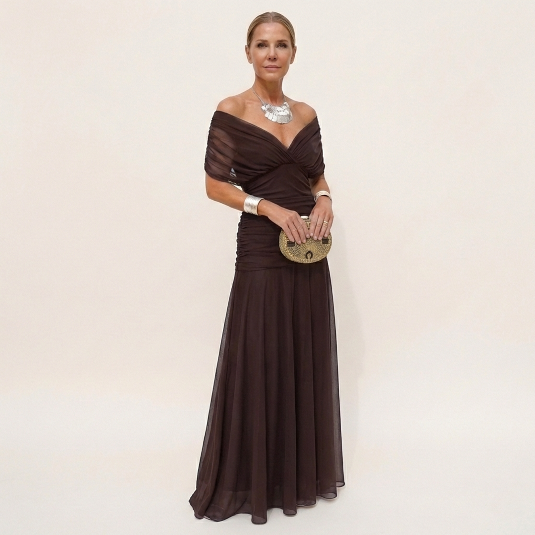 Model in a dark brown off-the-shoulder maxi dress with ruched bodice and flowing skirt, holding a gold clutch with silver jewelry. 2