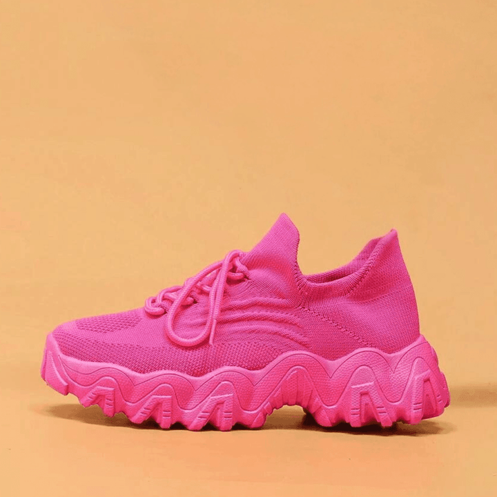 Side view of a single hot pink knit sneaker with a thick wavy sole on an orange backdrop. 22
