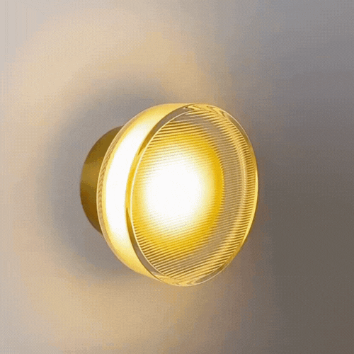 Round wall lamp with ribbed clear glass front and brass-toned backplate, casting a warm golden glow.