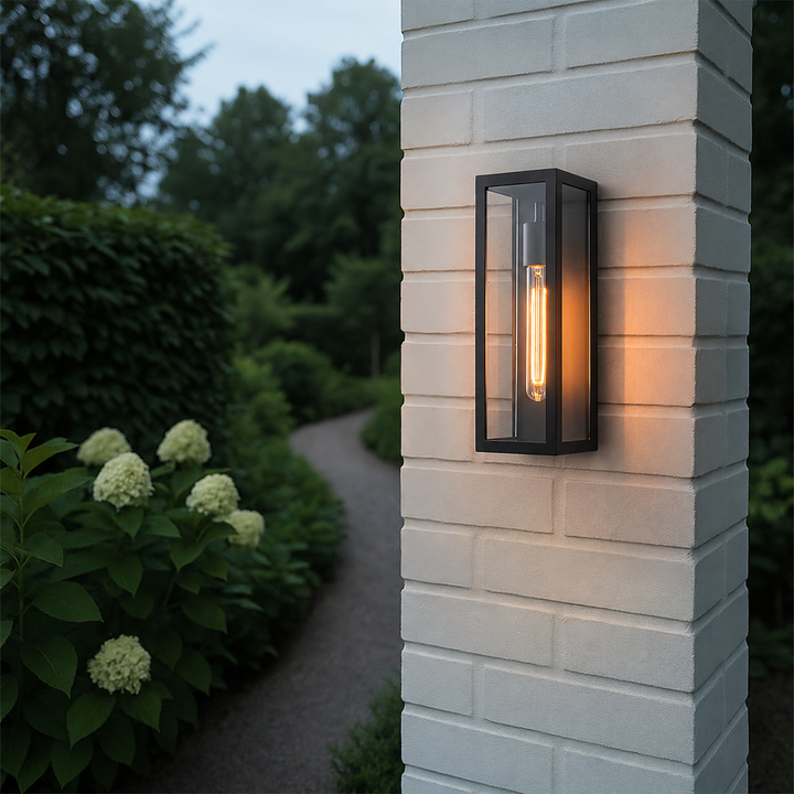 Black outdoor wall lantern with clear glass glowing on a white painted brick pillar beside a garden path.
