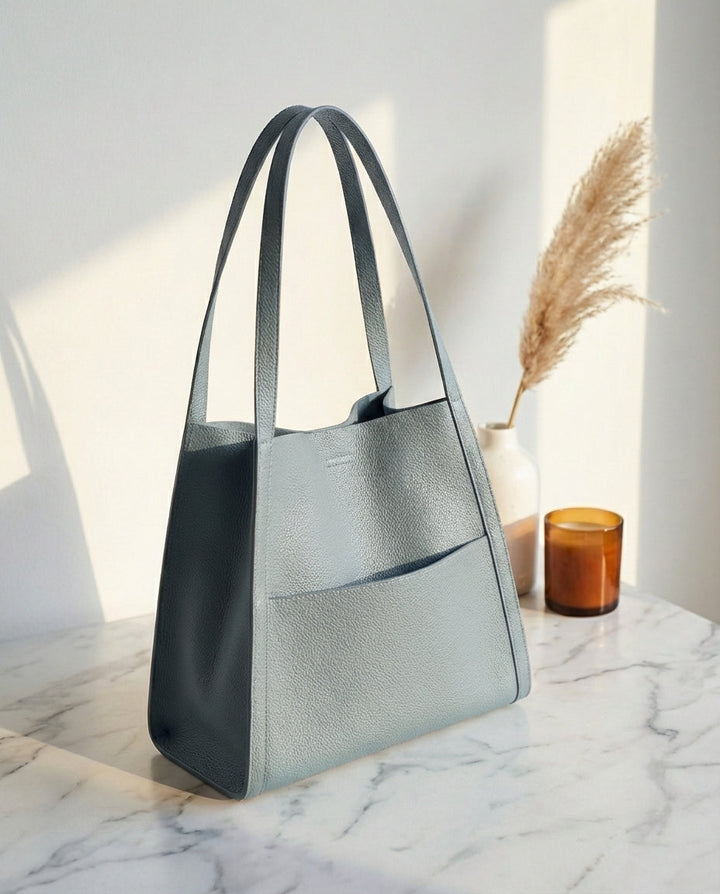 Light blue-gray pebbled leather tote with a front pocket, shown on a marble surface beside pampas grass and an amber candle.