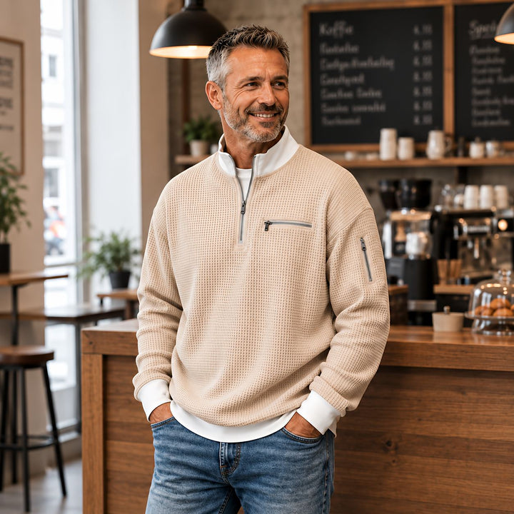 Man wearing a beige waffle knit quarter-zip sweater with a white collar, leaning on a wooden counter.
