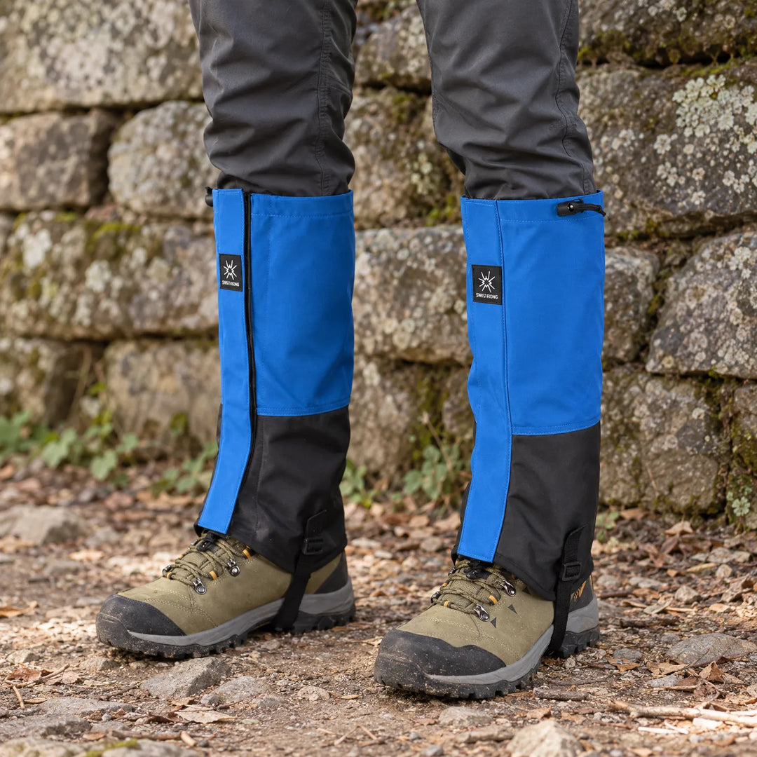 Blue and black gaiters worn over olive hiking boots, standing outdoors against a mossy stone wall with leaves on the ground. 1