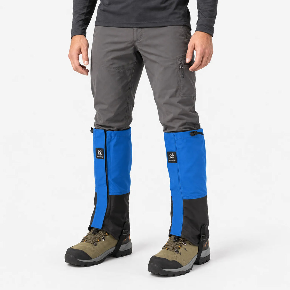 Man in gray pants wearing blue and black gaiters over hiking boots, three-quarter view on a plain white background. 2