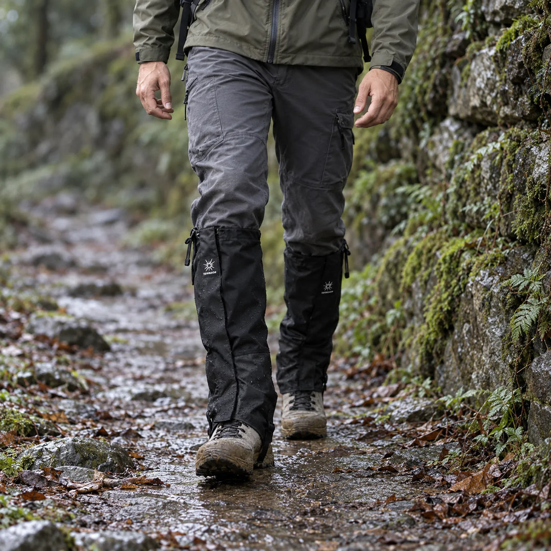 Hiker walking down a wet, leaf-covered forest trail in black gaiters speckled with water droplets and muddy boots. 3