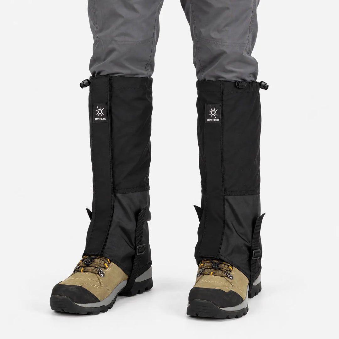Front view of all-black gaiters with drawcord tops and instep straps, worn over gray pants and tan hiking boots. 8