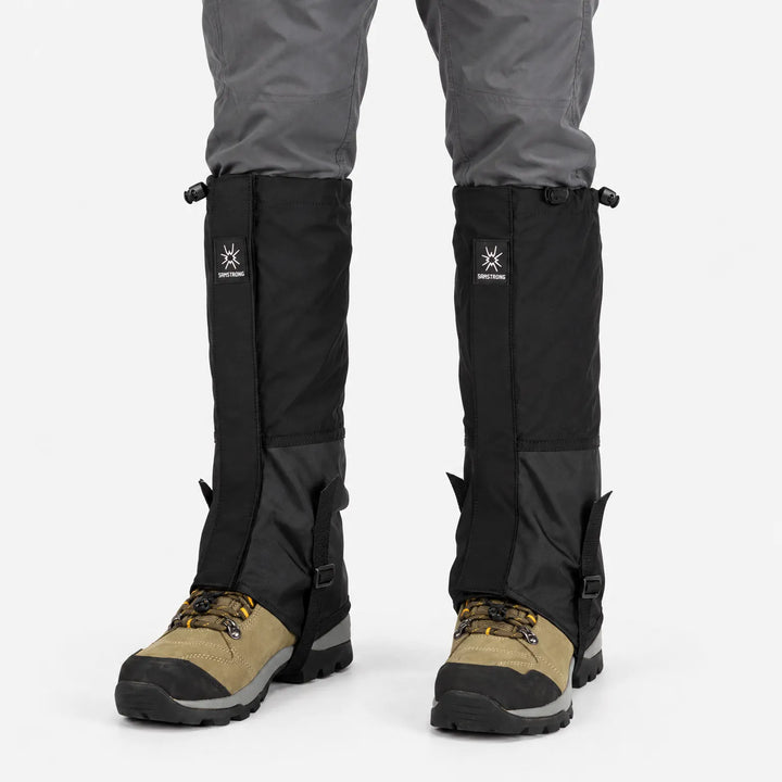 Front view of all-black gaiters with drawcord tops and instep straps, worn over gray pants and tan hiking boots. 8