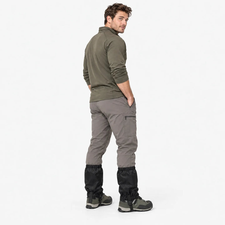 Man in an olive pullover and gray cargo pants, seen from behind, wearing black gaiters over hiking boots in a studio. 4