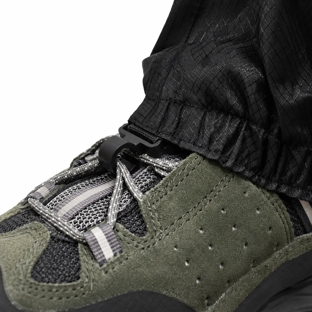 Close-up of the gaiter's lower hem with a black plastic hook clipped to the laces of an olive suede hiking shoe. 6