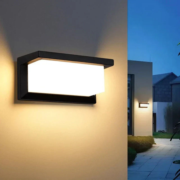Lit black wall light on a cream wall at dusk, with a second light and a paved path beyond. 3