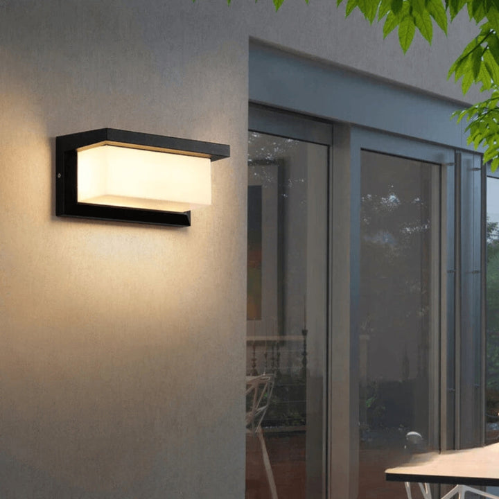 Black rectangular wall light glowing warm white on a gray stucco wall beside large glass doors, with green leaves overhead. 1