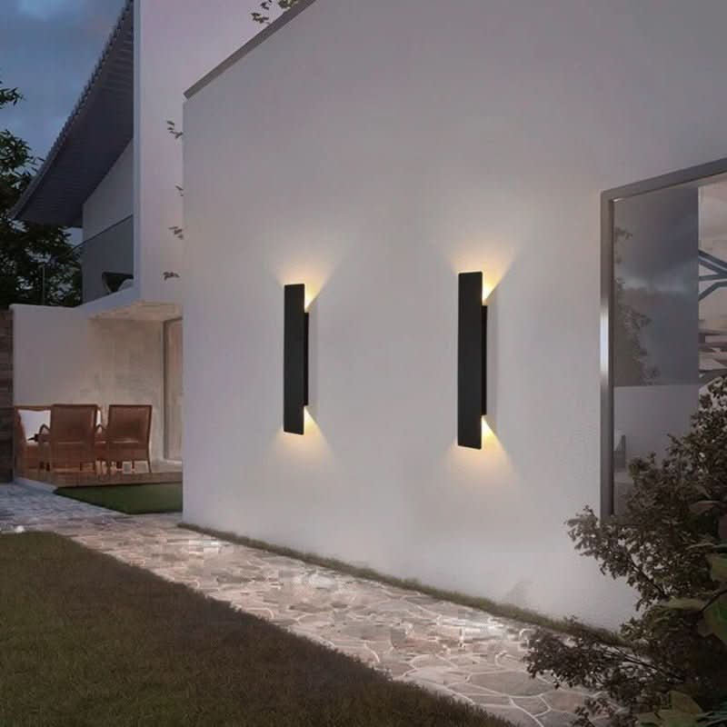 Pair of black vertical wall lights on a white exterior wall at night, lighting the wall above and below beside a stone path. 3