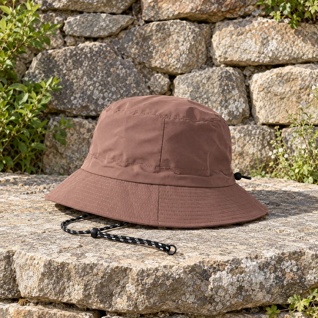 Brown bucket hat with a black-and-white cord resting on a sunlit stone ledge in front of a rough stone wall. 3