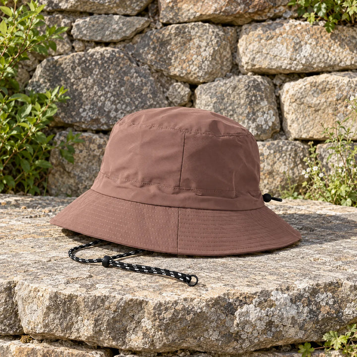 Brown bucket hat with a black-and-white cord resting on a sunlit stone ledge in front of a rough stone wall. 3