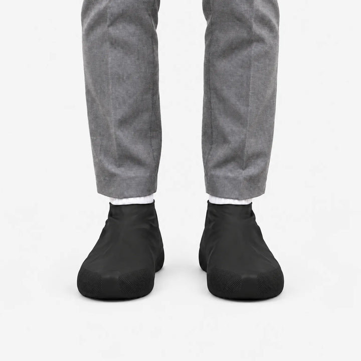 Front view of black shoe covers worn with gray trousers and white socks on a plain white background. 4
