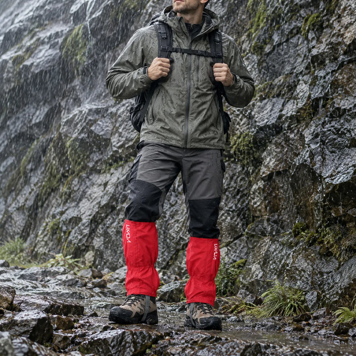 Hiker in a rain jacket and backpack stands on a wet rocky trail beside a rock wall, wearing red gaiters over boots. 1