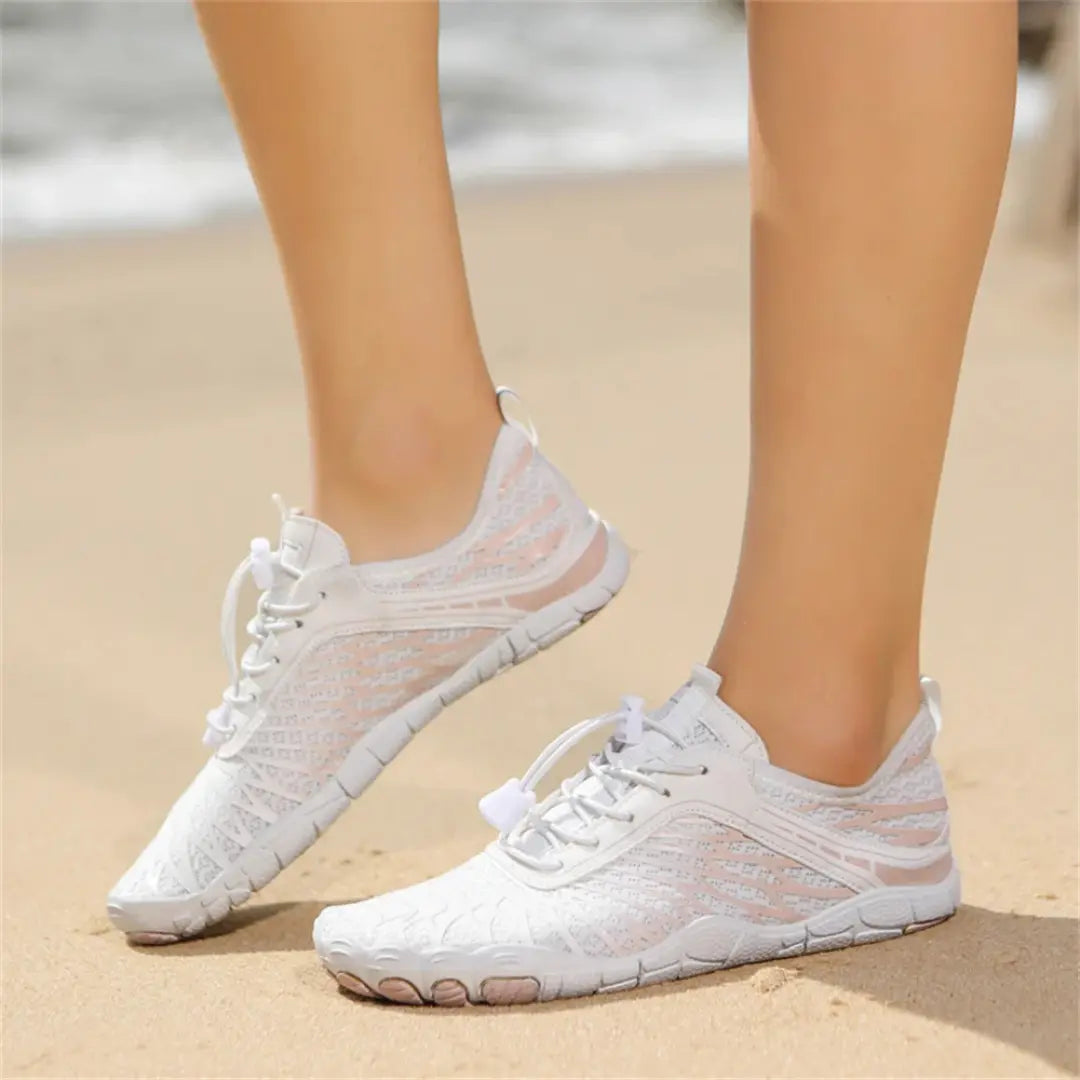 White barefoot shoes with pink details worn on a sandy beach, with soft waves blurred in the background.