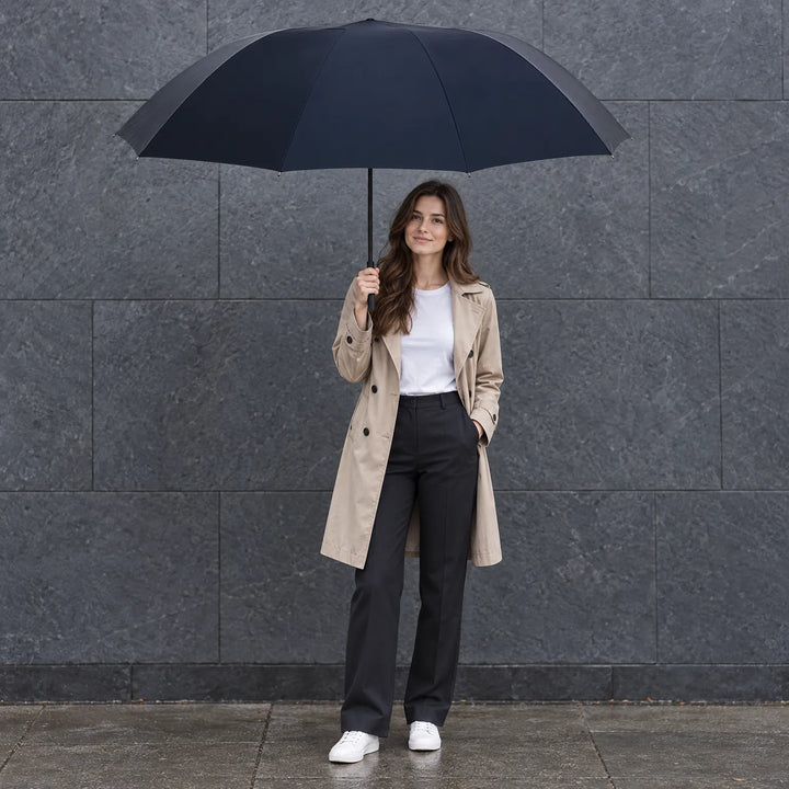 Woman in a beige trench coat and white sneakers holds a large navy umbrella while standing before a slate wall on wet pavement. 1