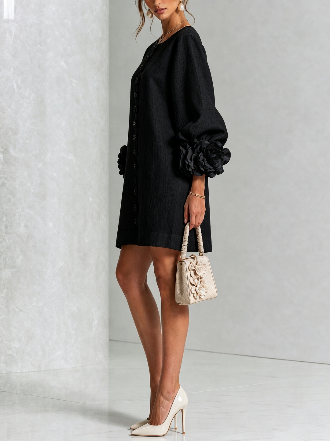 Black dress on a model in side view, showing the puffed sleeve with its large rose trim, worn with cream heels and a handbag. 22