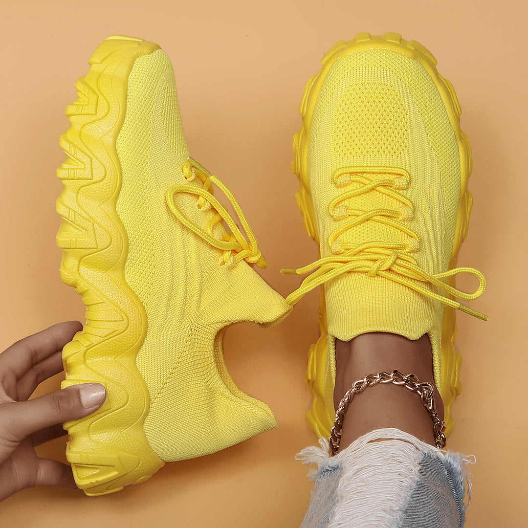 Overhead view of bright yellow knit lace-up sneakers with chunky matching soles, one held in hand and one worn. 47