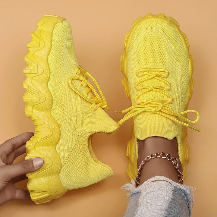 Overhead view of bright yellow knit lace-up sneakers with chunky matching soles, one held in hand and one worn. 47