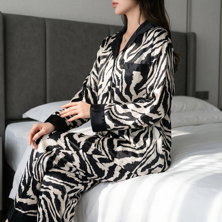 Woman sitting on the edge of a bed in a long-sleeve zebra-print satin pajama set with black collar, cuffs and chest pocket. 1