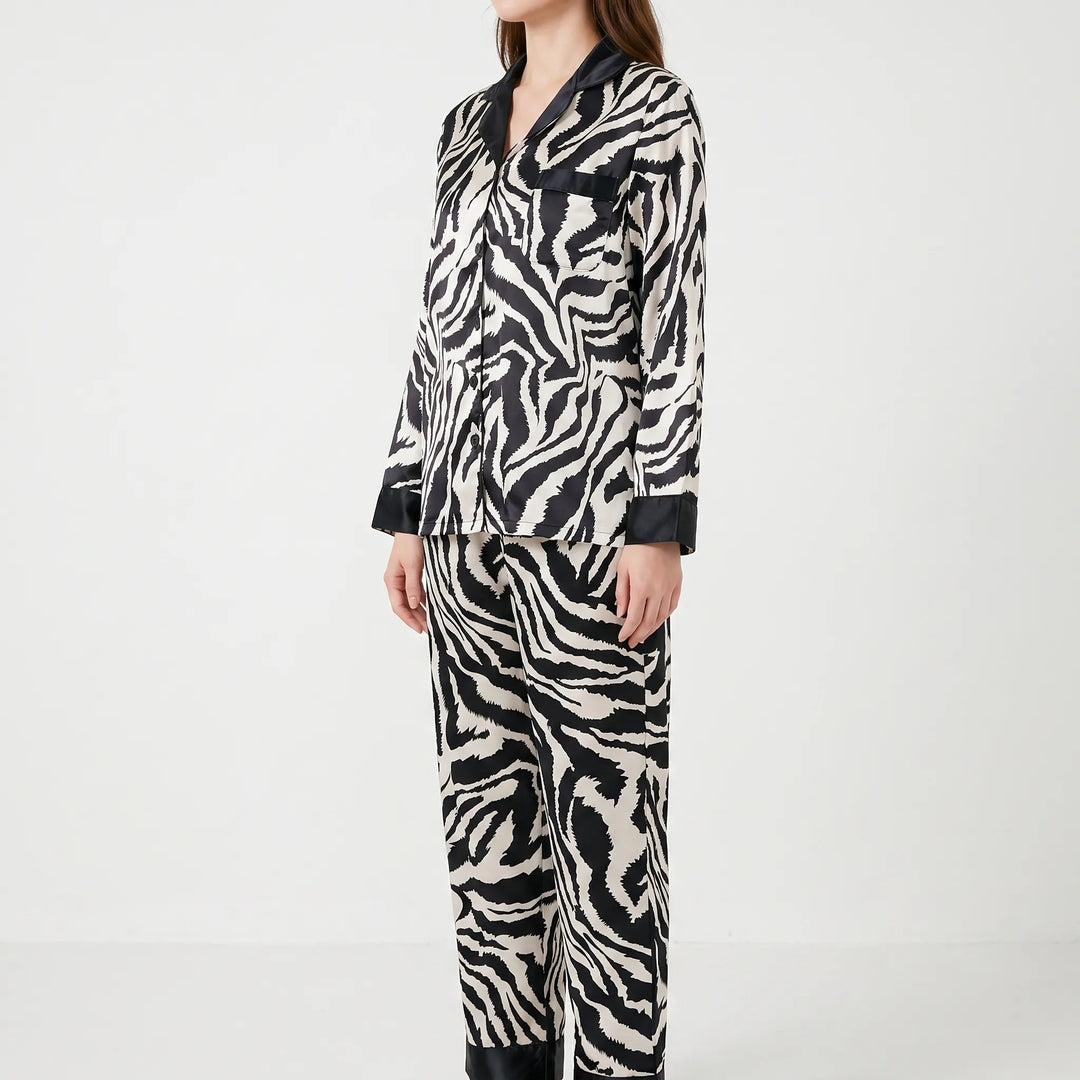 Three-quarter side view of a model standing in the long-sleeve zebra-print satin pajama set with black trim and a chest pocket. 3