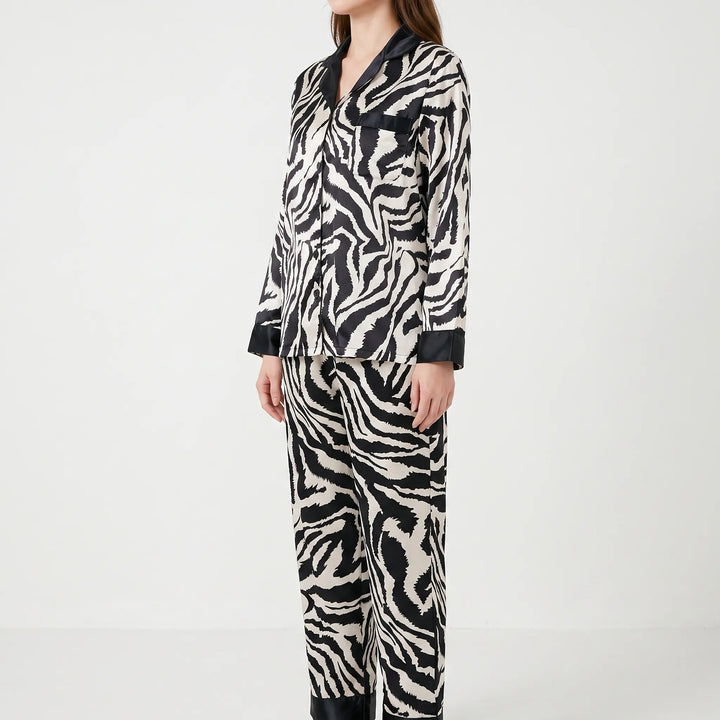 Three-quarter side view of a model standing in the long-sleeve zebra-print satin pajama set with black trim and a chest pocket. 3