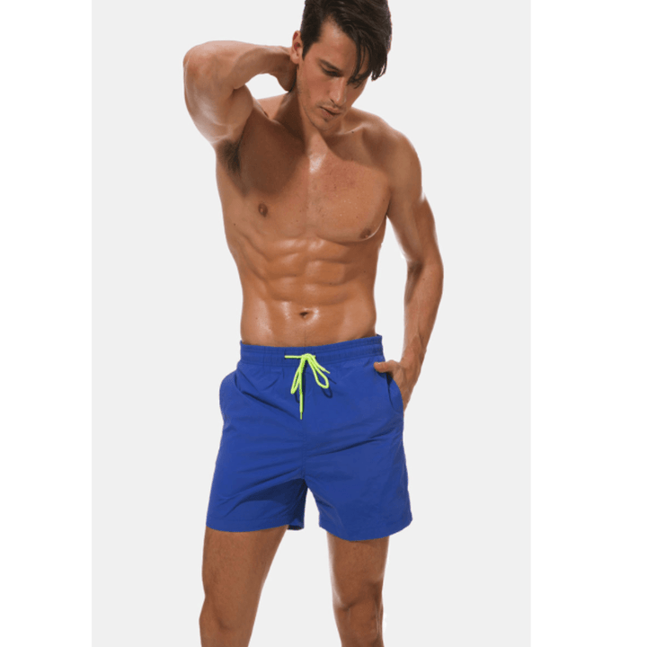 Model with wet skin posing in royal blue swim trunks with a neon yellow drawstring, one hand in a pocket. 4
