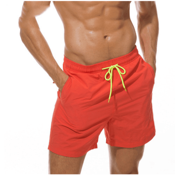 Coral red swim trunks with a neon yellow drawstring, worn by a model with both hands in the side pockets. 6